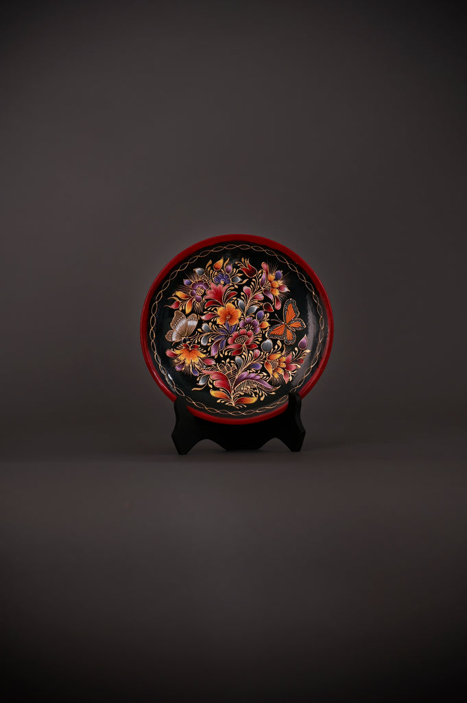 Hand-painted Mexican decorative plate featuring vibrant flowers and monarch butterflies on a black background, crafted using the traditional laca perfilada technique with a red rim and displayed on a black stand.