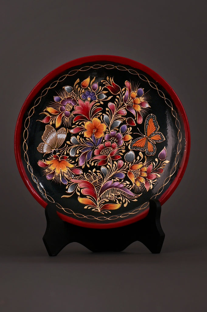 Hand-painted Mexican decorative plate featuring vibrant flowers and monarch butterflies on a black background, crafted using the traditional laca perfilada technique with a red rim and displayed on a black stand.