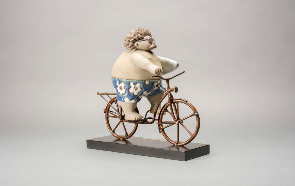 "Handmade ceramic sculpture of a surfer riding a bicycle with a surfboard by Rodo Padilla – Mexican folk art surf decor" 27x9x24 cm (approx. 10.63 x 3.54 x 9.45 inches)